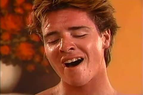 A close-up shot of a sweaty, smooth young man with his eyes closed and mouth open in an expression of intense pleasure.