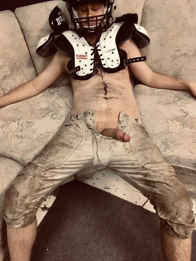 A man wearing a football helmet and shoulder pads lies back with his penis exposed through his dirty football pants.