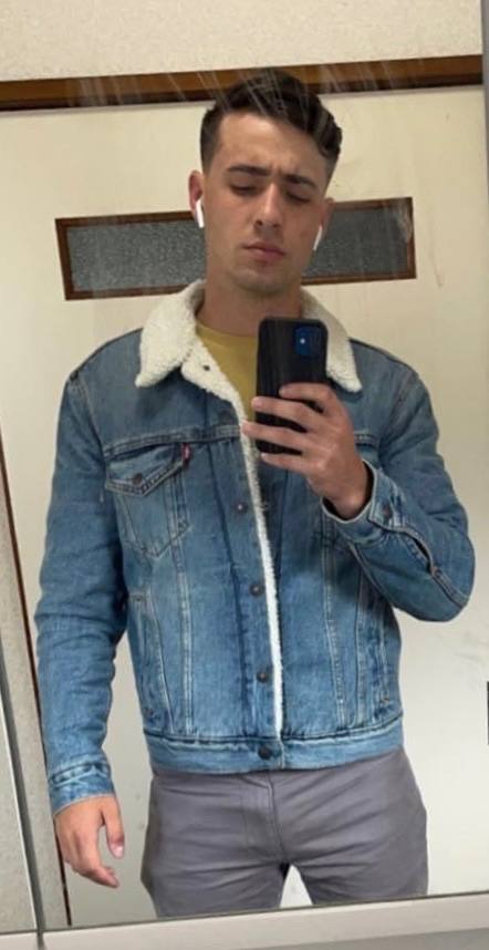 A young man with a buzzcut takes a mirror selfie while wearing a shearling-collared denim jacket and tight grey pants.