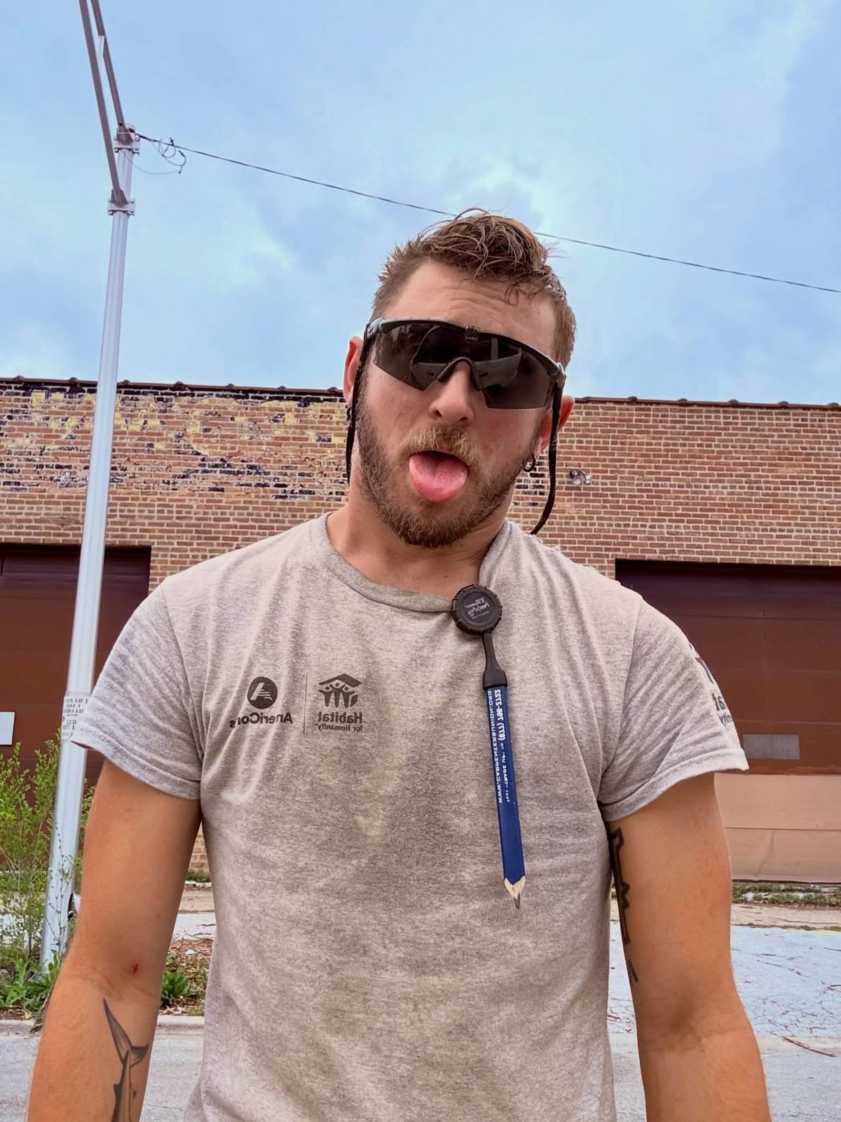 A bearded man with tattoos wears a grey construction t-shirt and safety sunglasses while sticking his tongue out in an outdoor setting.