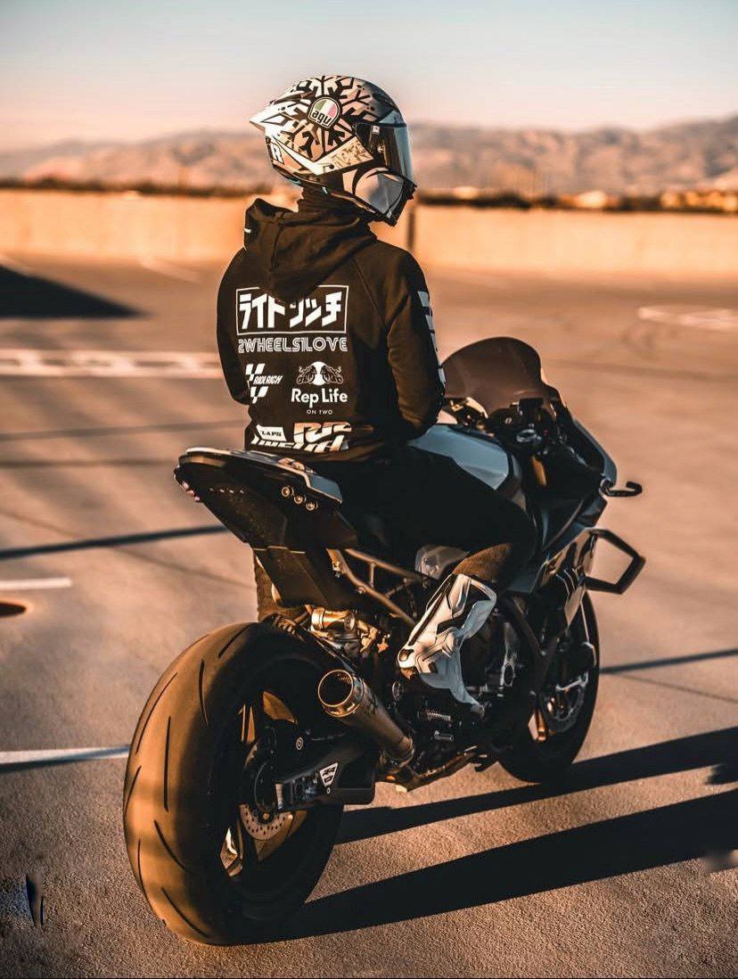 A person wearing a full-face racing helmet, a black hoodie, and white motorcycle boots sits on a sportbike in an outdoor parking lot at sunset.