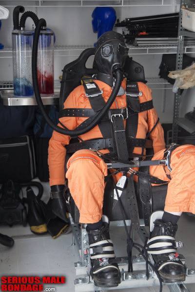 A person wearing an orange jumpsuit and gas mask is strapped securely into a mechanical restraint chair.