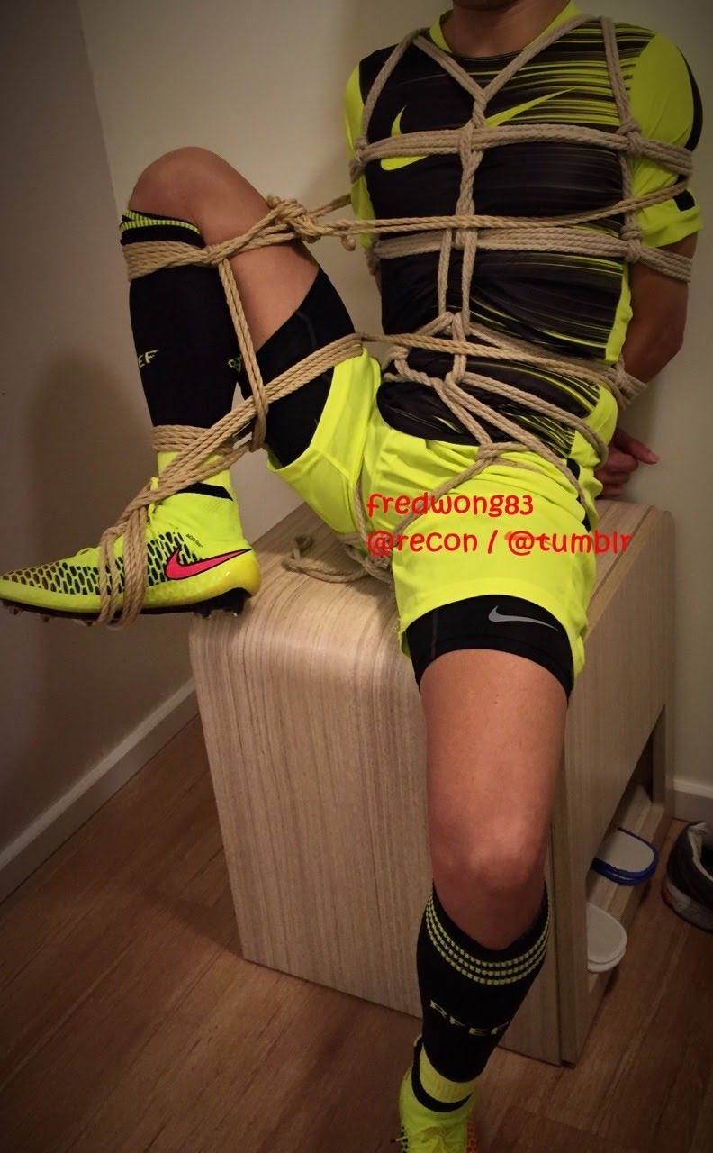 A man dressed in a neon yellow soccer kit and cleats is bound in detailed rope shibari while sitting on a wooden box.