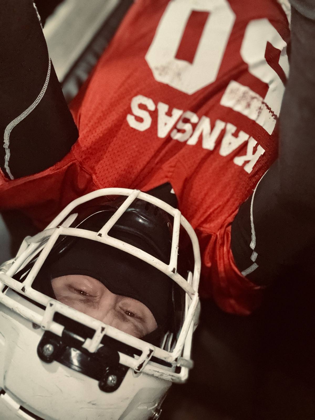 A man wearing a red Kansas jersey, black compression gear, a balaclava, and a football helmet lies down in a gym setting.