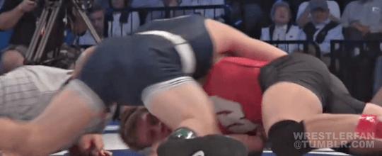 Two young men in wrestling singlets engage in a grappling match on an indoor mat.
