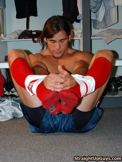 A shirtless muscular jock sits on the floor stretching while holding his feet together in long red sports socks.