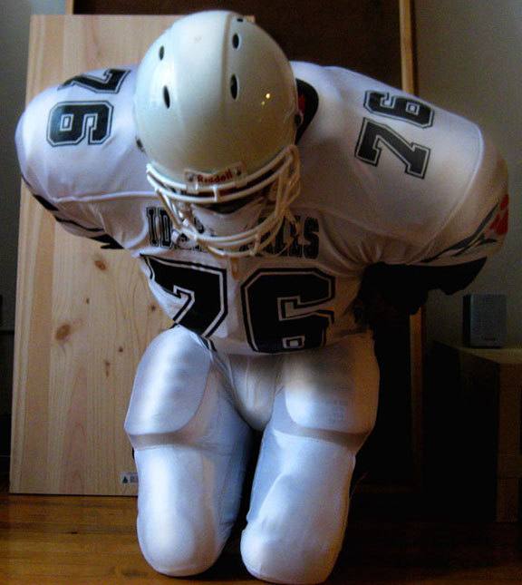 A white football uniform with a helmet, jersey, and padded pants is displayed in an indoor setting.