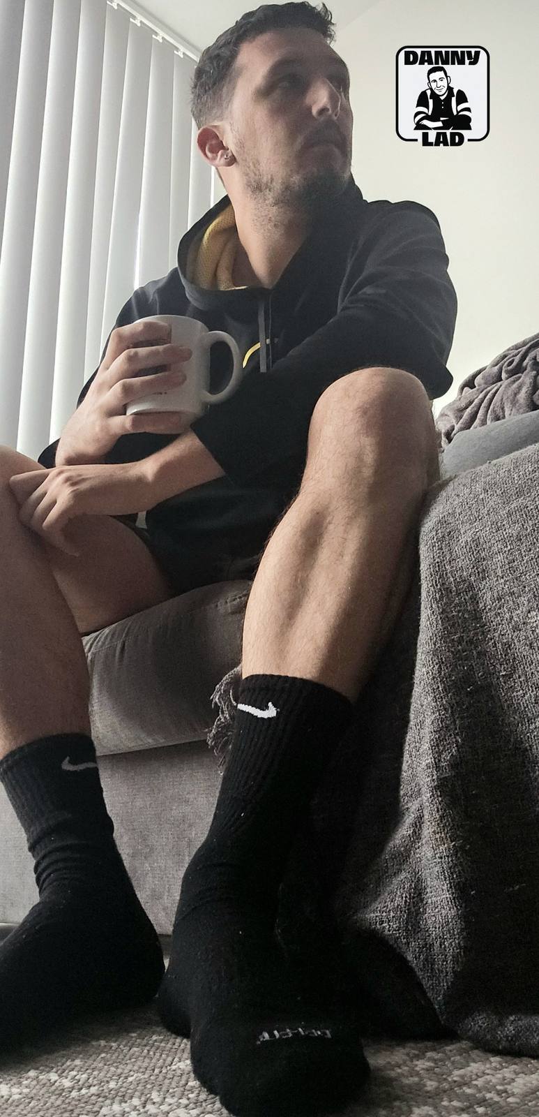 A bearded man in a black hoodie and shorts sits on a sofa holding a mug, showcasing his muscular legs and black Nike crew socks.