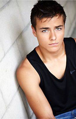 A young man in a black tank top and jeans poses indoors.