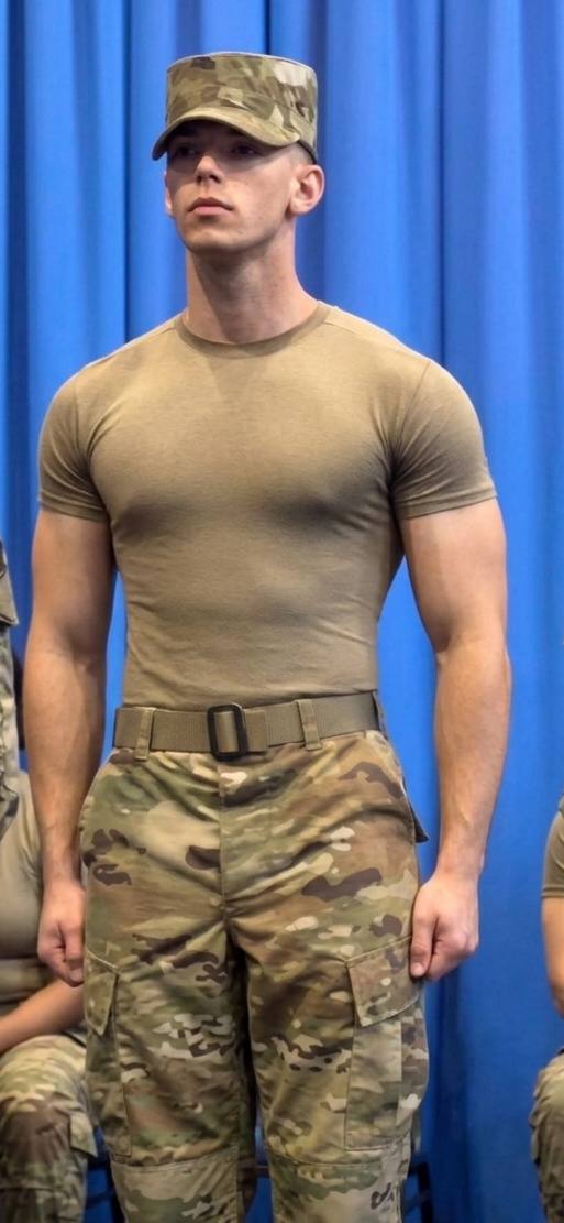 A muscular young man stands at attention wearing a tight tan undershirt and camouflage military fatigues against a blue backdrop.