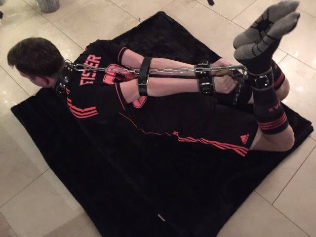 A man wearing a black football kit lies prone on a black blanket while restrained with a leather collar, cuffs, and metal chains.