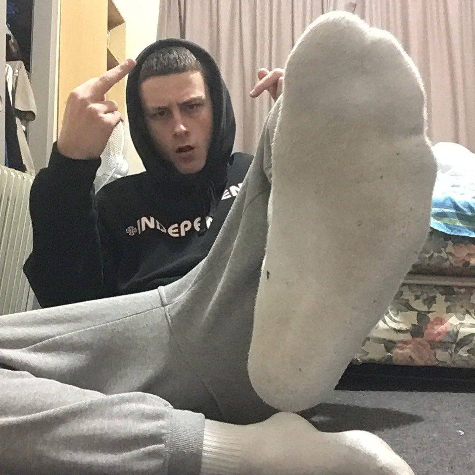 A young man wearing a black hoodie and grey sweatpants sits on the floor and extends his white-socked foot toward the camera while making a gesture.