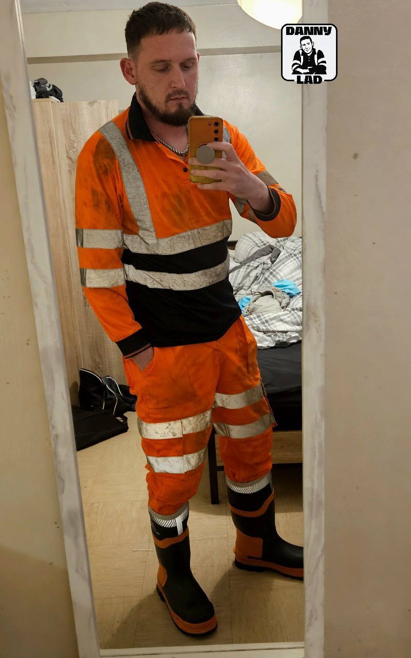 A bearded man with a buzzcut takes a mirror selfie wearing a full orange hi-vis construction uniform and rubber boots.
