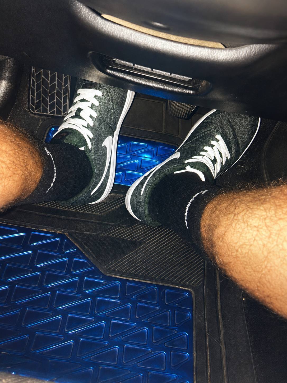 A POV shot shows a person with hairy legs wearing black socks and green Nike sneakers while sitting in the driver's seat of a car.