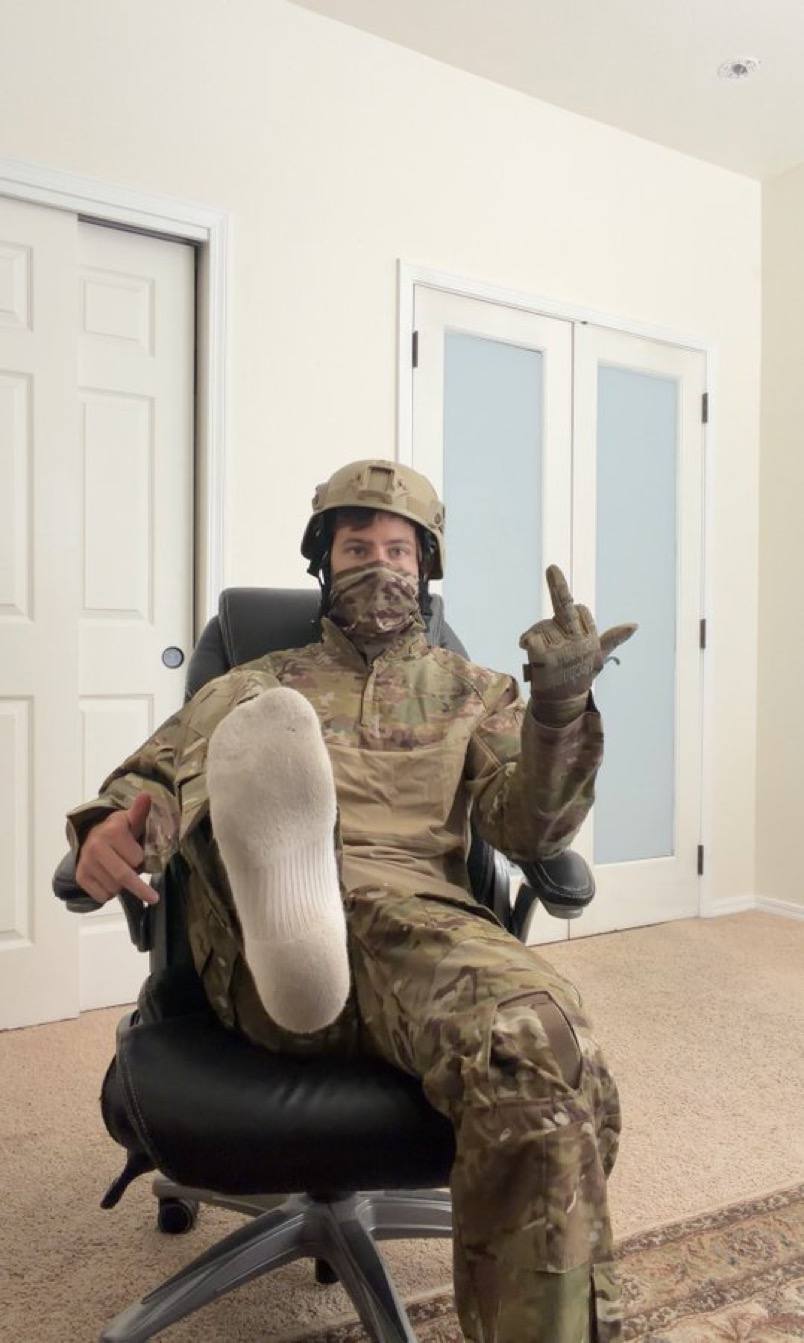 A young man in full military camouflage gear and a tactical helmet sits in a chair, presenting the sole of his white socked foot to the camera.