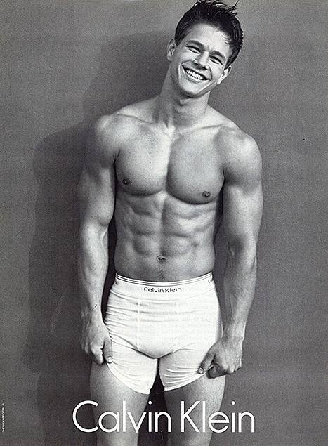 A black and white studio portrait of a muscular, smooth-chested man wearing white Calvin Klein boxer briefs.