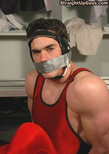 A muscular man wearing a red wrestling singlet and ear guards is gagged with duct tape in a locker room setting.