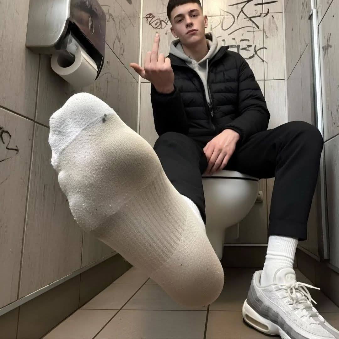 A young man sits on a toilet in a public restroom with one socked foot extended.