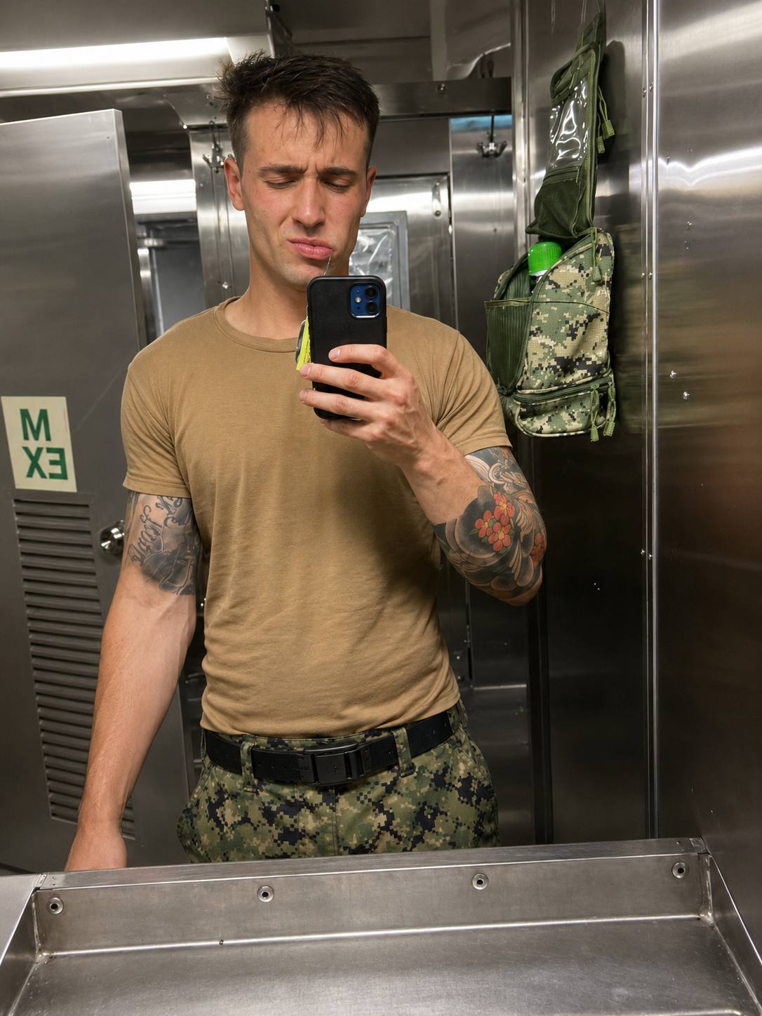 A tattooed man in a tan military undershirt and digital camo pants takes a mirror selfie in a stainless steel locker room.