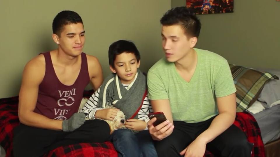 Two young men and a child sit on a bed with a small dog while looking at a phone.