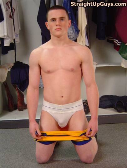 A muscular young man with a buzzcut kneels in a locker room wearing only a white jockstrap while holding his athletic shorts down.