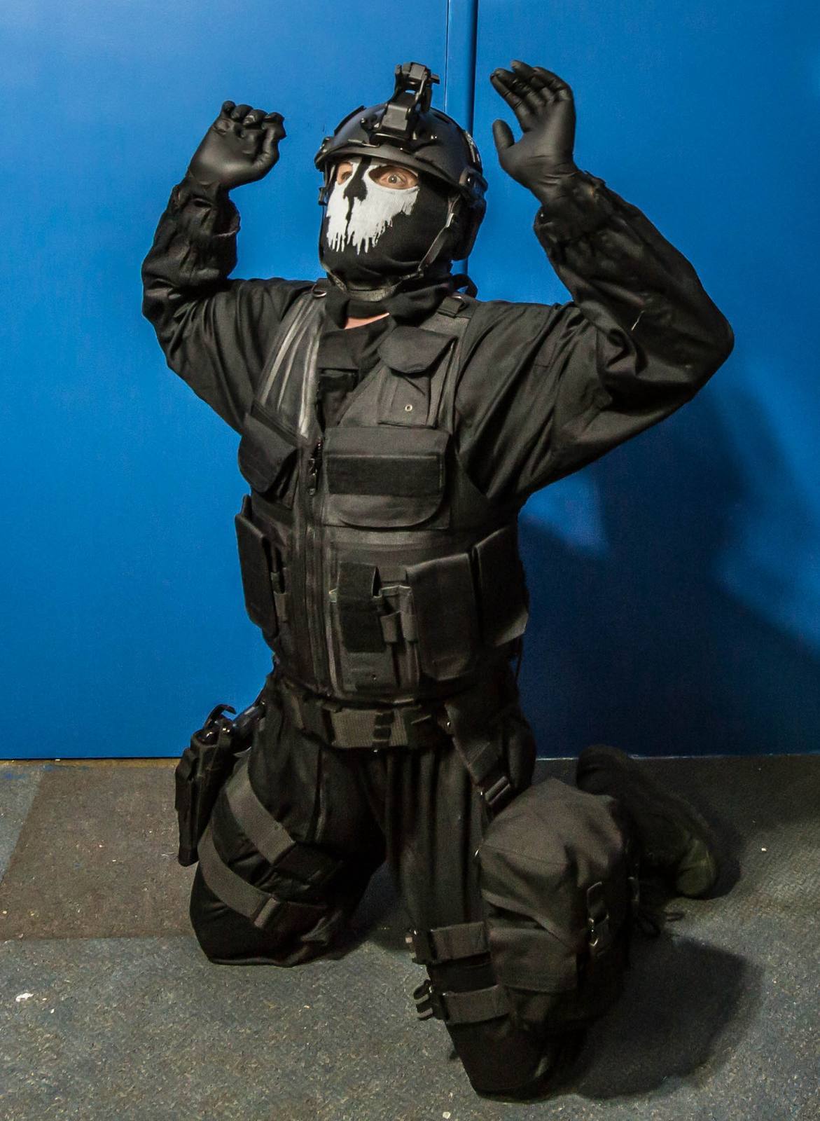 A man dressed in a complete black tactical military uniform with a skull mask kneels submissively with his hands up.