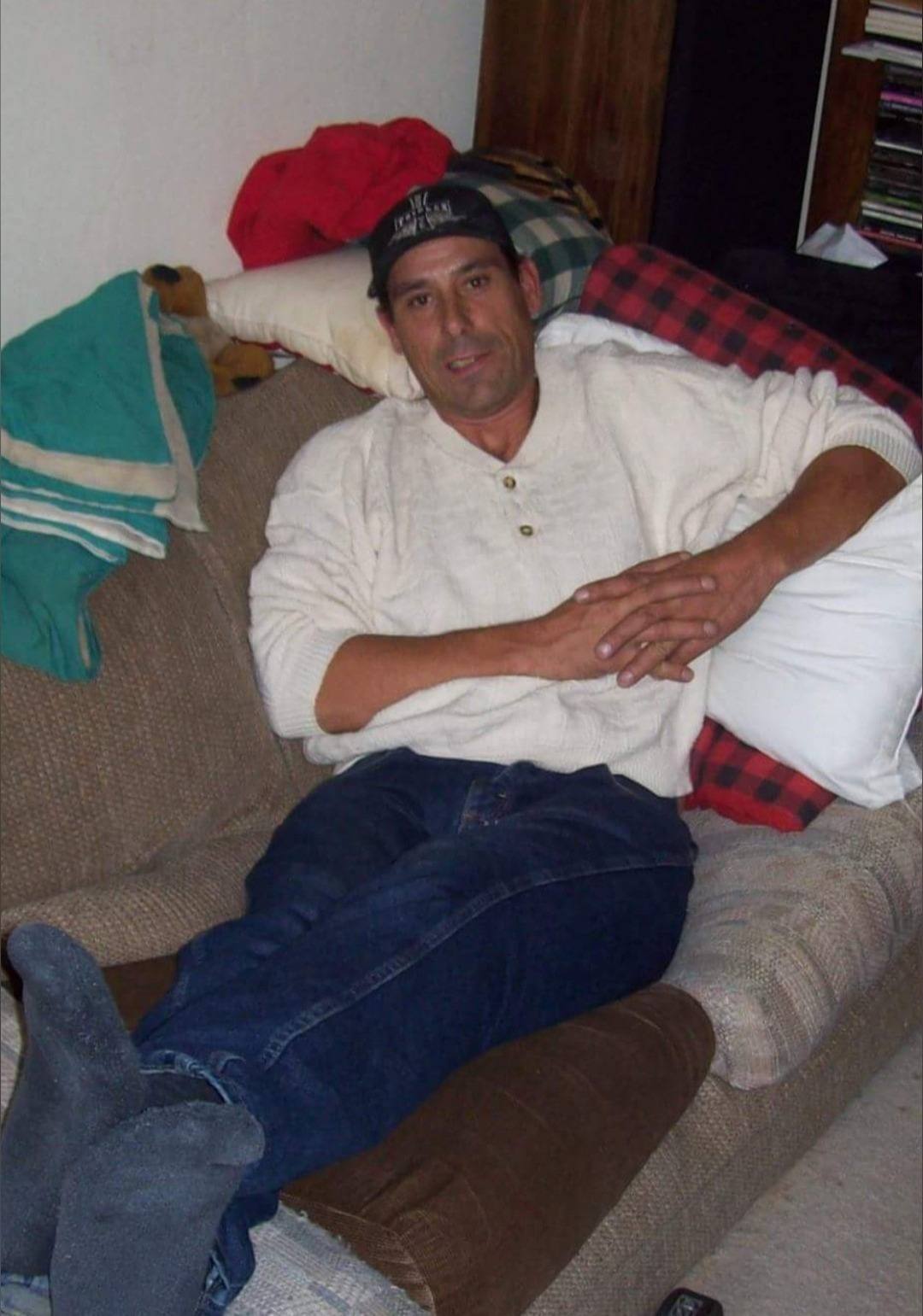 A man lounges on a sofa wearing a shirt, jeans, socks, and a cap.