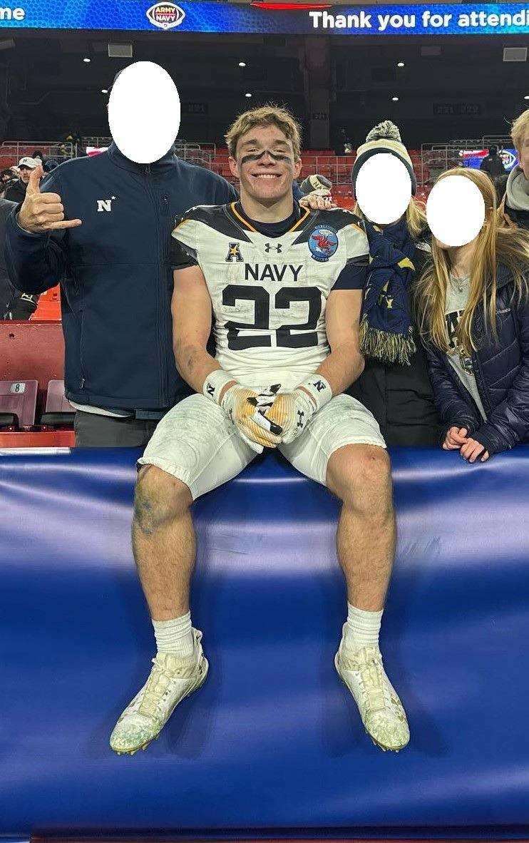 A muscular young football player in a dirty Navy jersey and white pants sits on a stadium barrier after a game.