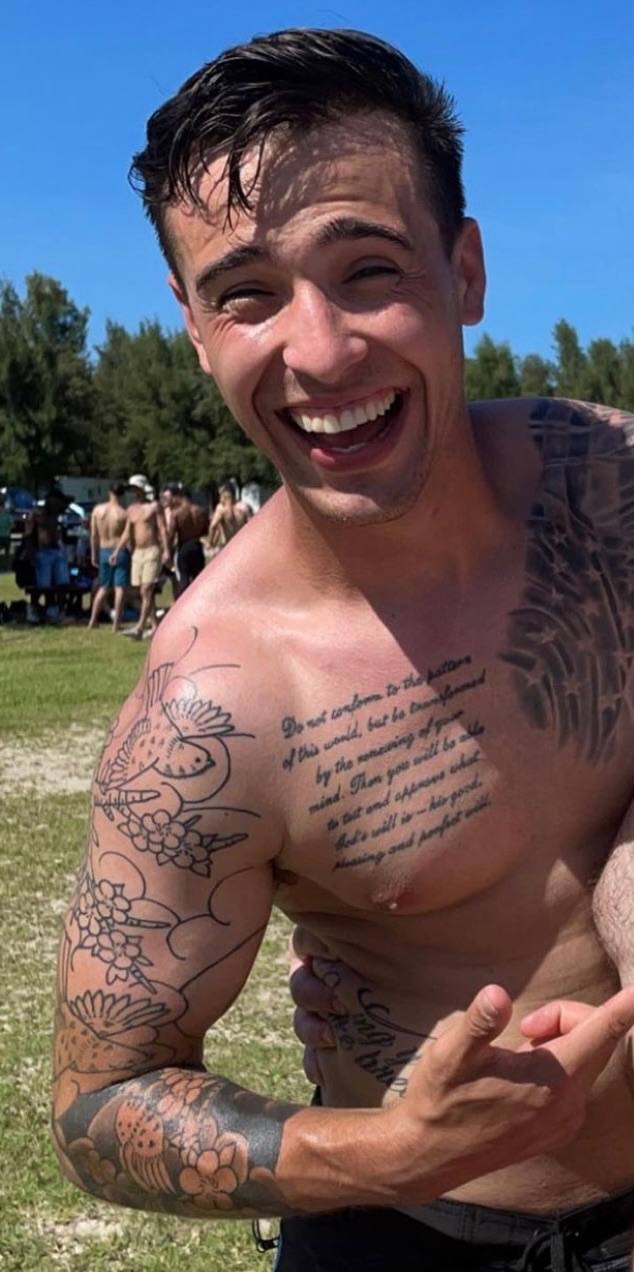 A smiling, topless man with heavy arm and chest tattoos stands in an outdoor park setting.