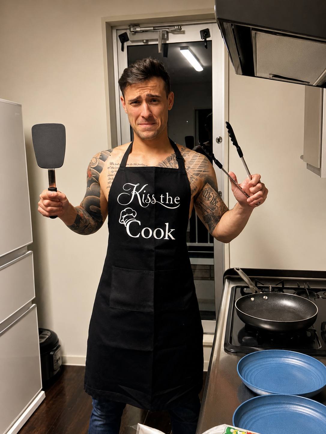 A shirtless, tattooed twunk stands in a kitchen wearing a black Kiss the Cook apron while holding a spatula and tongs.