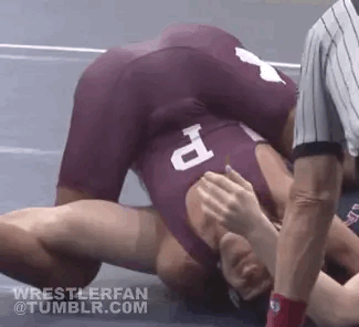 A wrestler wearing a maroon singlet pins his opponent to the mat during a grappling match.
