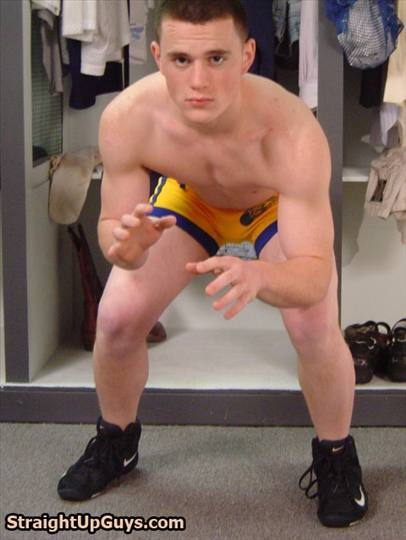 A muscular young man with a buzzcut poses in a wrestling stance wearing a yellow and blue singlet and black sneakers in a locker room.
