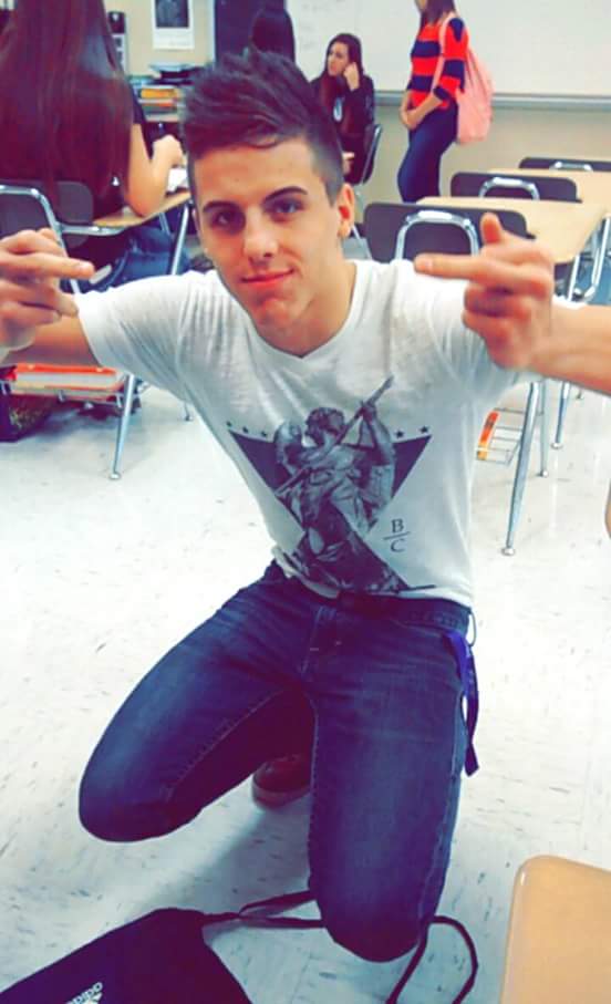 A young man kneels on the floor in a classroom, wearing a t-shirt and jeans.