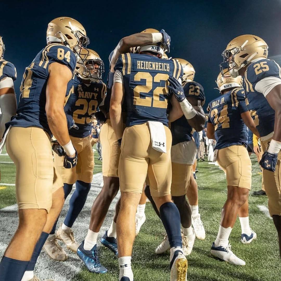 A group of muscular football players in tight uniforms huddle together on the field at night, their spandex pants accentuating their athletic builds.