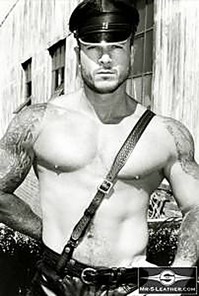 A muscular, tattooed man wears a black leather Muir cap and cross-chest leather strap outdoors against a warehouse background.