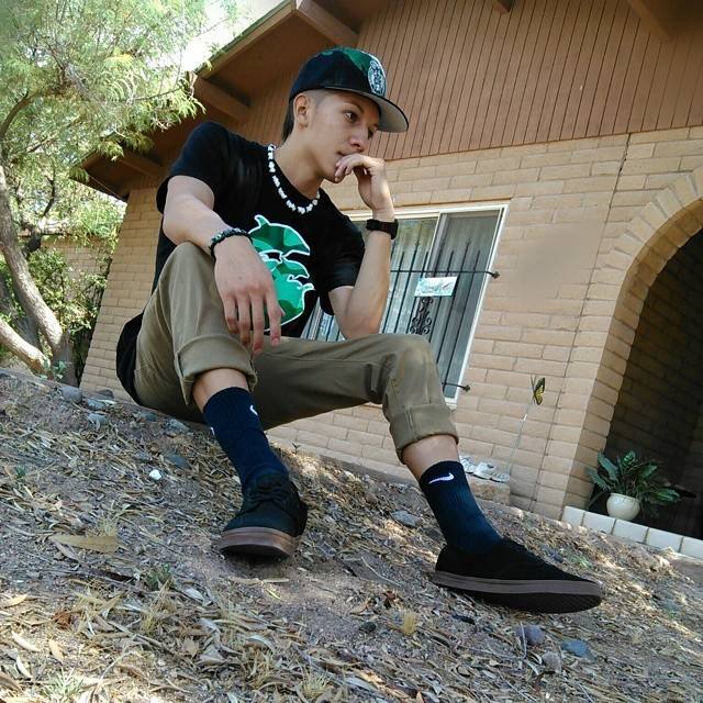 A young man sits outdoors on a dirt slope, wearing a cap, t-shirt, and sneakers.