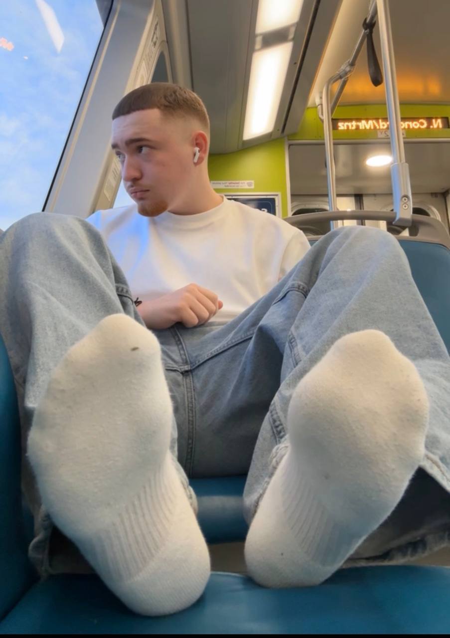 A young man with a buzzcut sits on a train with his feet in white socks propped up toward the camera.