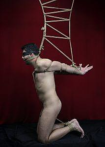 A nude man kneels blindfolded while bound in intricate rope shibari suspension bondage against a dark red backdrop.