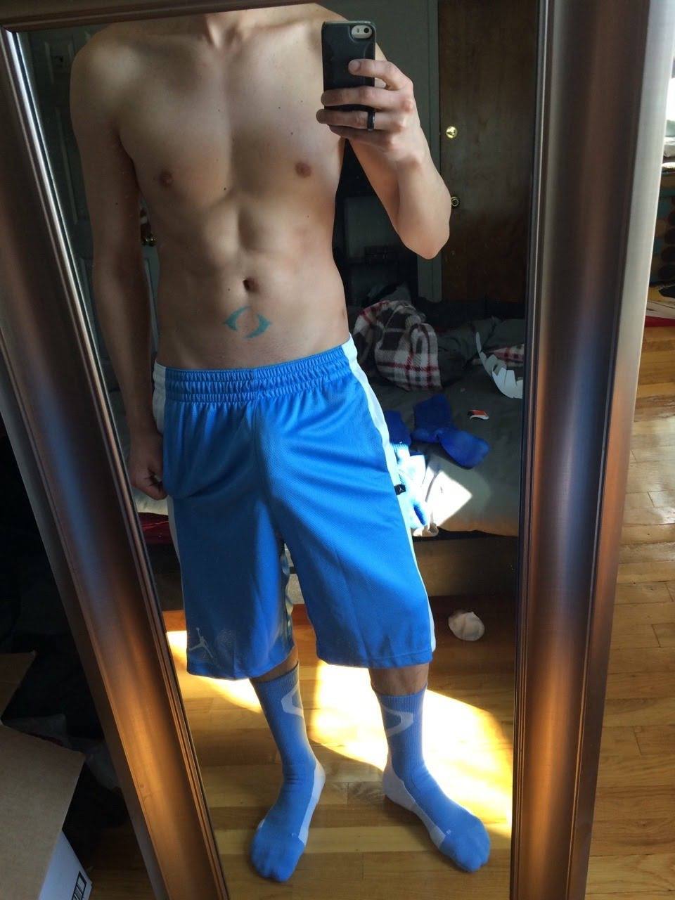 A shirtless twunk shows off his bulge in light blue gym shorts and matching socks while taking a mirror selfie.