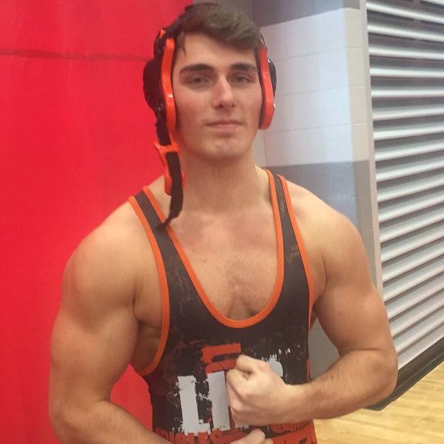 A muscular young man in a patterned wrestling singlet and protective headgear poses in a gym.