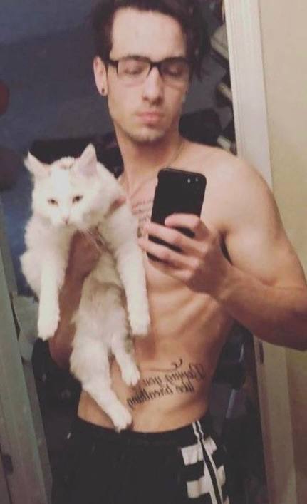 A shirtless young man with glasses and abdominal tattoos takes a mirror selfie while holding a large white cat.