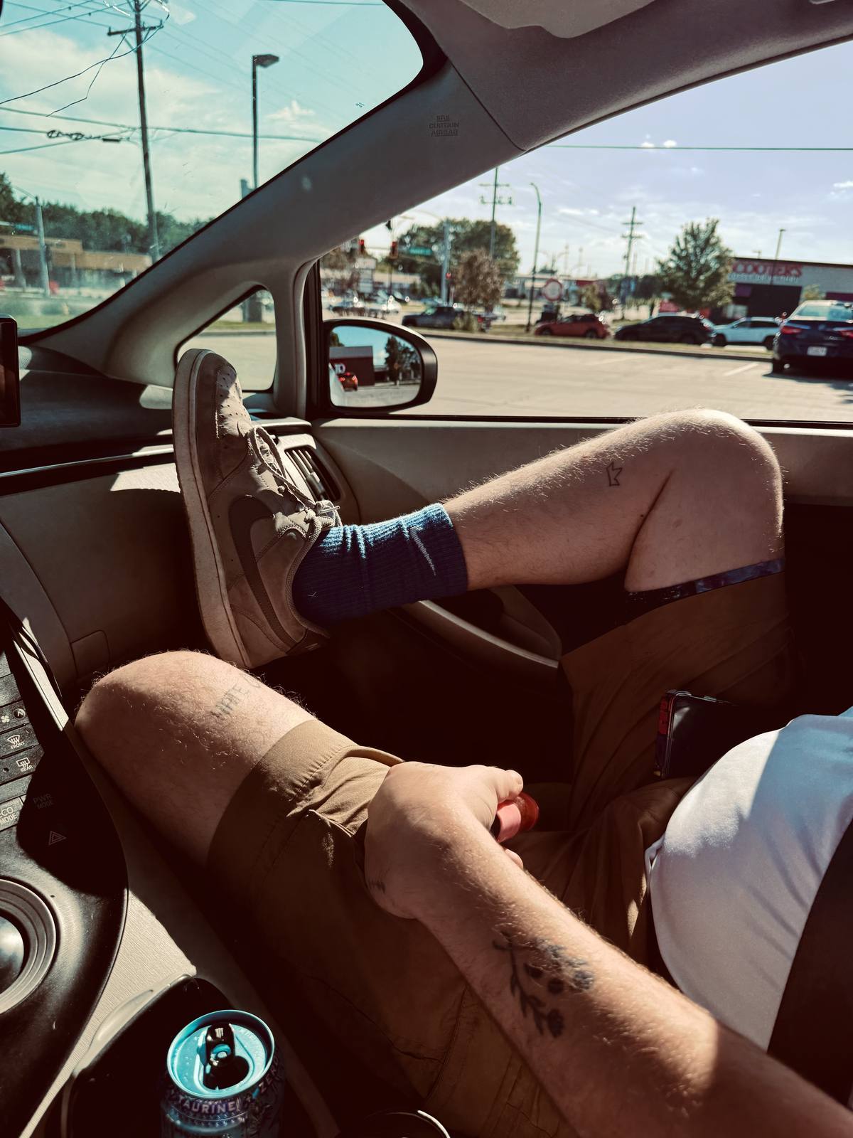 A man relaxes in the passenger seat of a car with his hairy, tattooed legs propped up, wearing blue tube socks and sneakers.