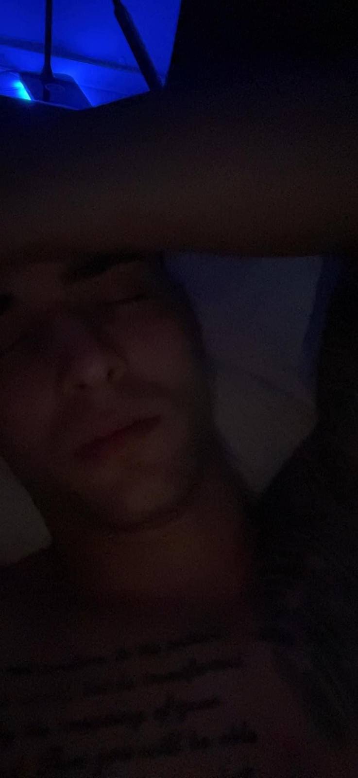 A close-up shot of a young, shirtless man with a chest tattoo lying down with his eyes closed in a dimly lit room.