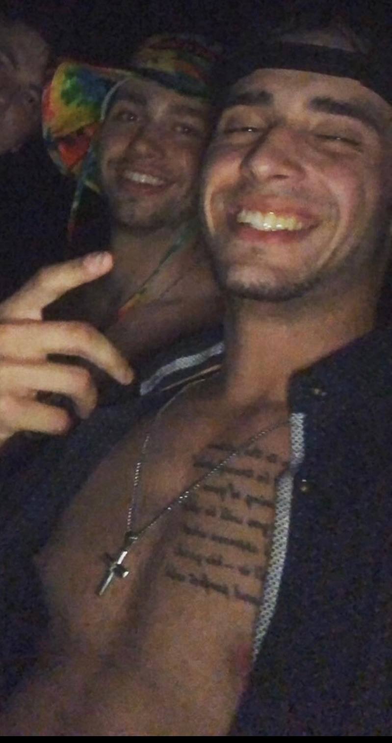 A young man with a script tattoo on his chest smiles for a selfie with friends in a dark outdoor setting.