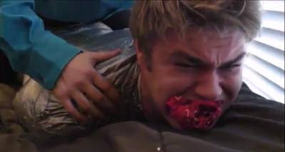 A man wrapped tightly in silver duct tape with a red cloth gag in his mouth lies prone on a bed while another person rests a hand on his back.