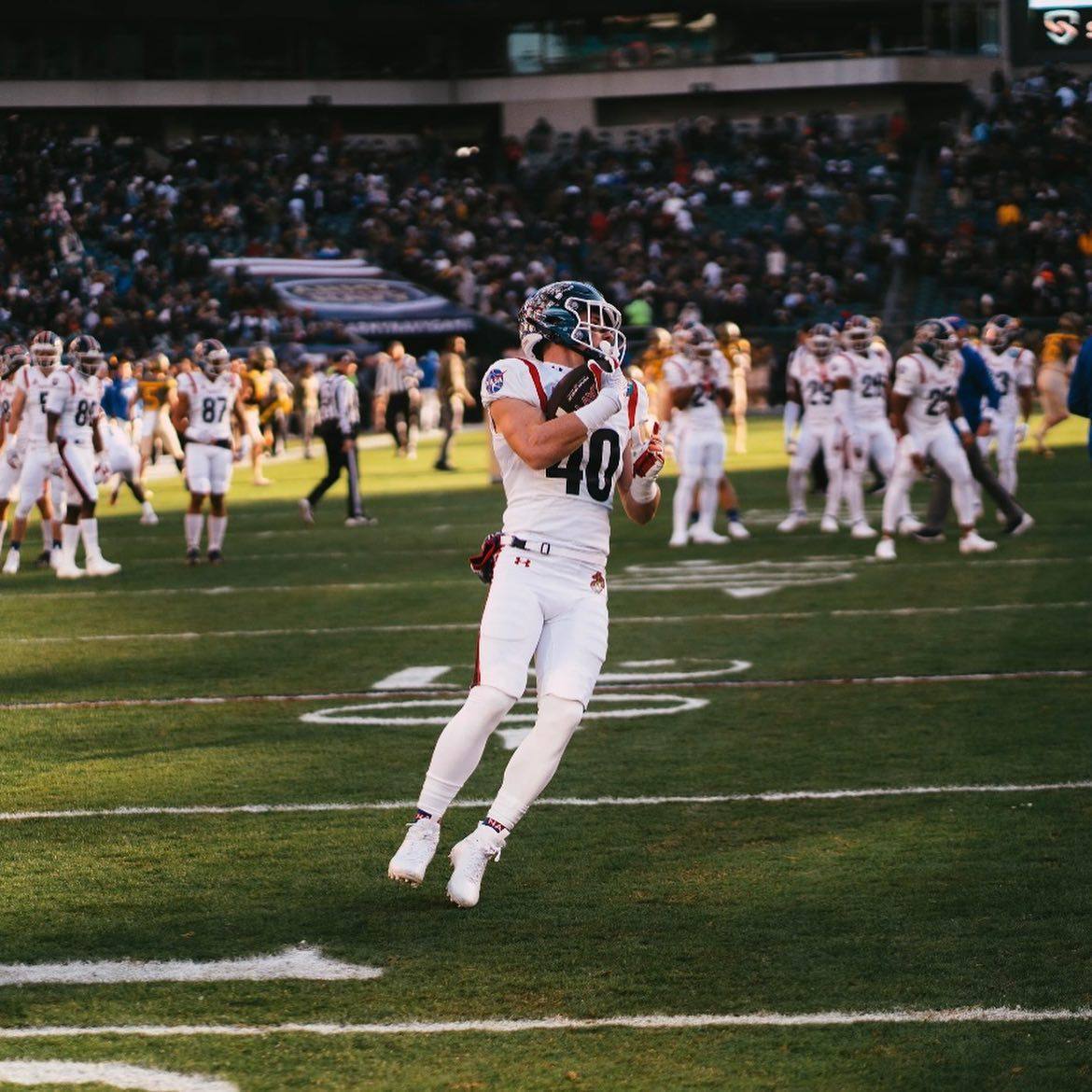 A muscular football player in a full white uniform catches a ball on a stadium field.