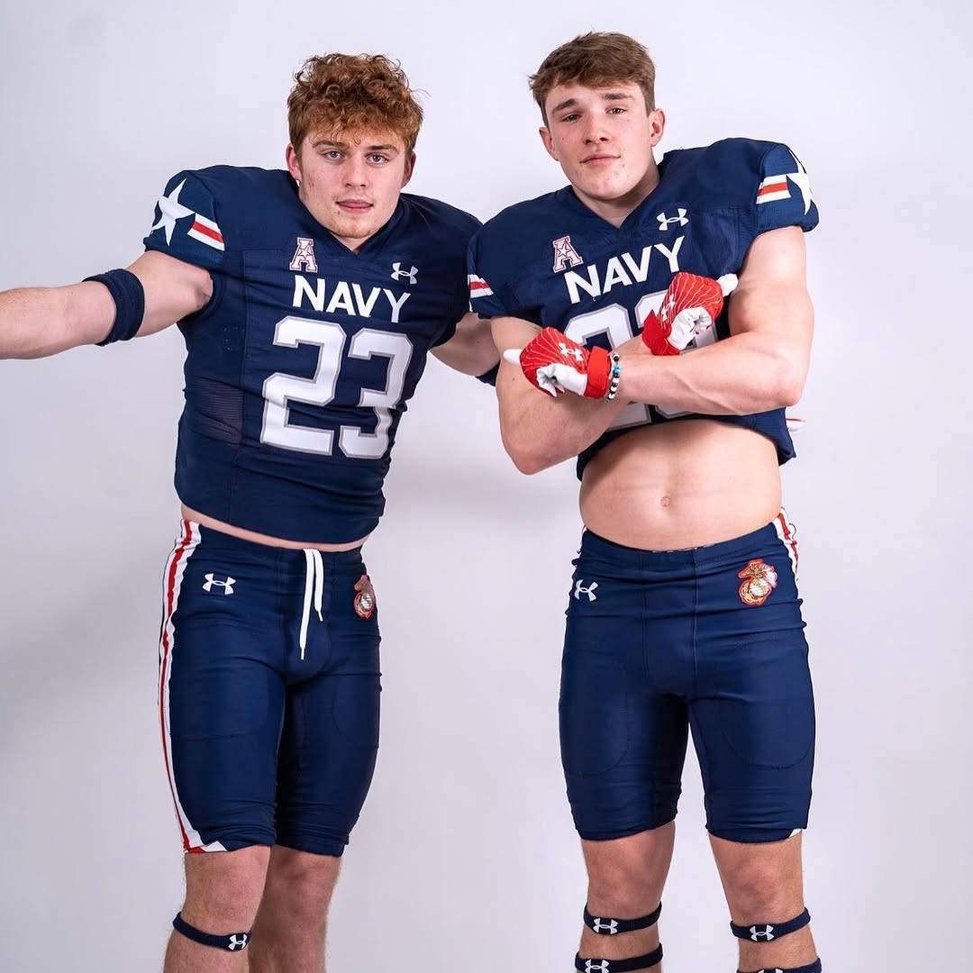 Two athletic young men pose in full Navy football kits, with one showing off his defined abs.
