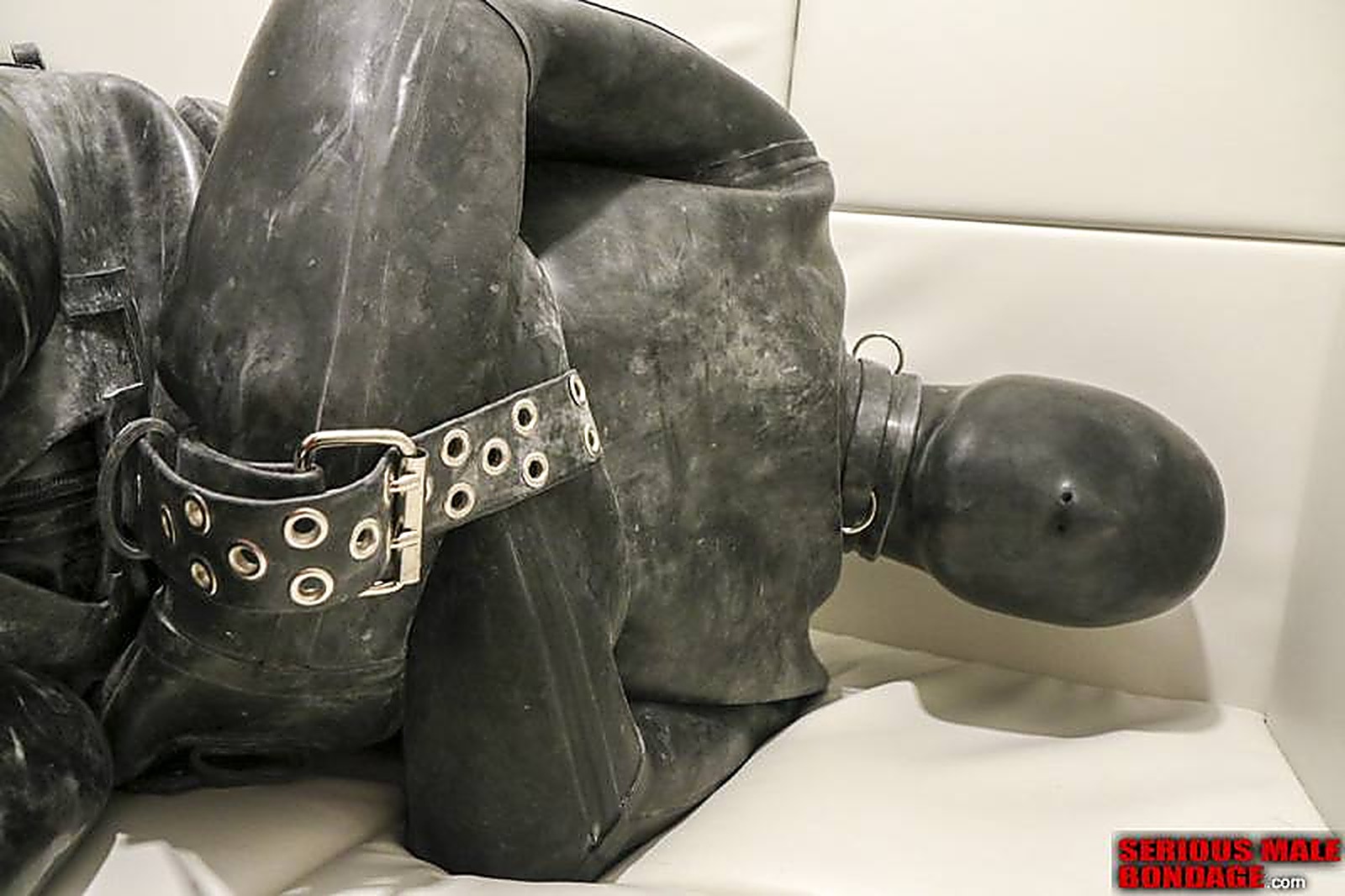 A person restrained in a full black rubber suit lies on a padded white bench.