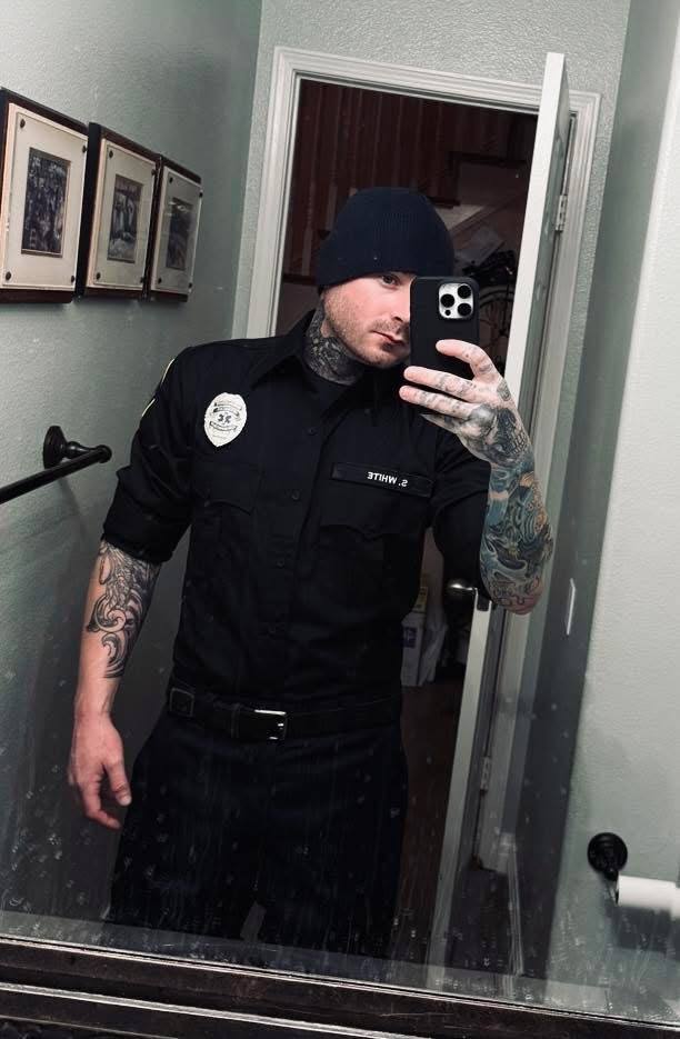 A heavily tattooed man in a black cop uniform and beanie takes a mirror selfie.