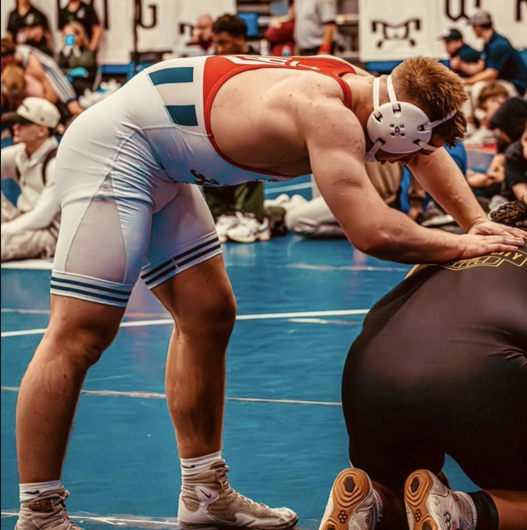 A muscular wrestler wearing a tight white and red singlet and headgear leans over his opponent on a blue wrestling mat.